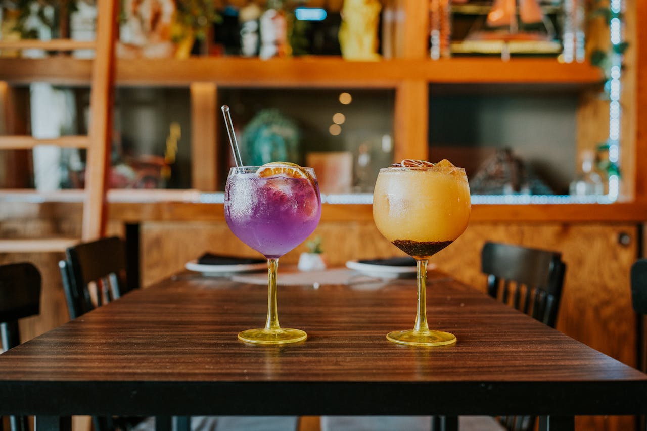 why-choose-us Two vibrant cocktails, purple and orange, on a rustic wooden table indoors. Perfect for a lively night out.